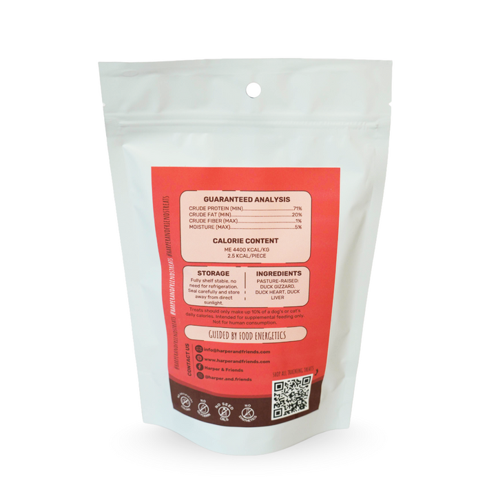 A stand-up resealable pouch of pet treats with a large orange and white label showing guaranteed analysis, calorie content, storage, ingredients, and a QR code. The pouch is photographed on a clean white background.