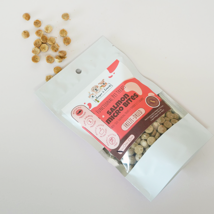 A stand-up pouch of raw organic pet treats labeled salmon micro bites, placed on a white background with several loose beige treats scattered to the upper left.