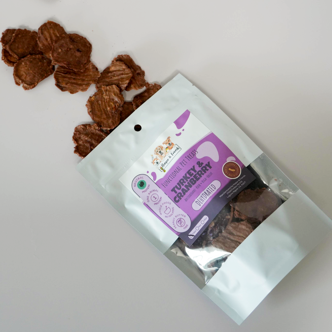 A white stand-up pouch of dog treats with a purple and white label, shown on a white background with several brown treat pieces spilled beside it. The label reads &